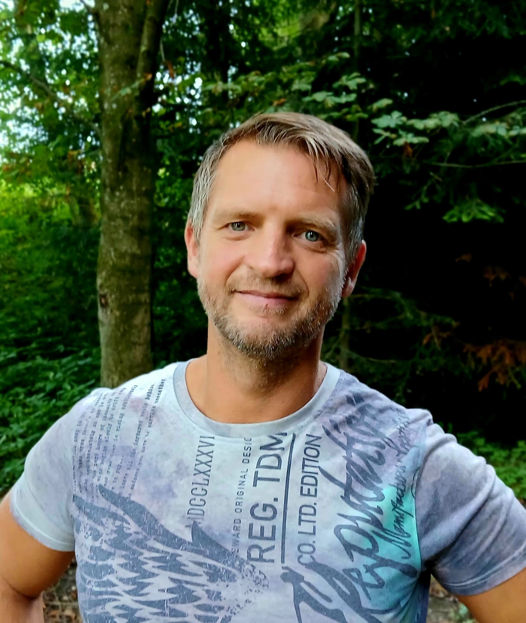 Mittelalter Mann mit leichtem Bart und bedrucktem T-Shirt lächelt in einem üppigen grünen Wald.