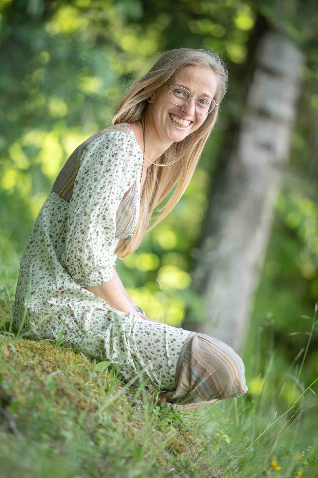 Lächelnde Frau mit Brille und blonden Haaren sitzt im geblümten Kleid auf einer Waldwiese.
