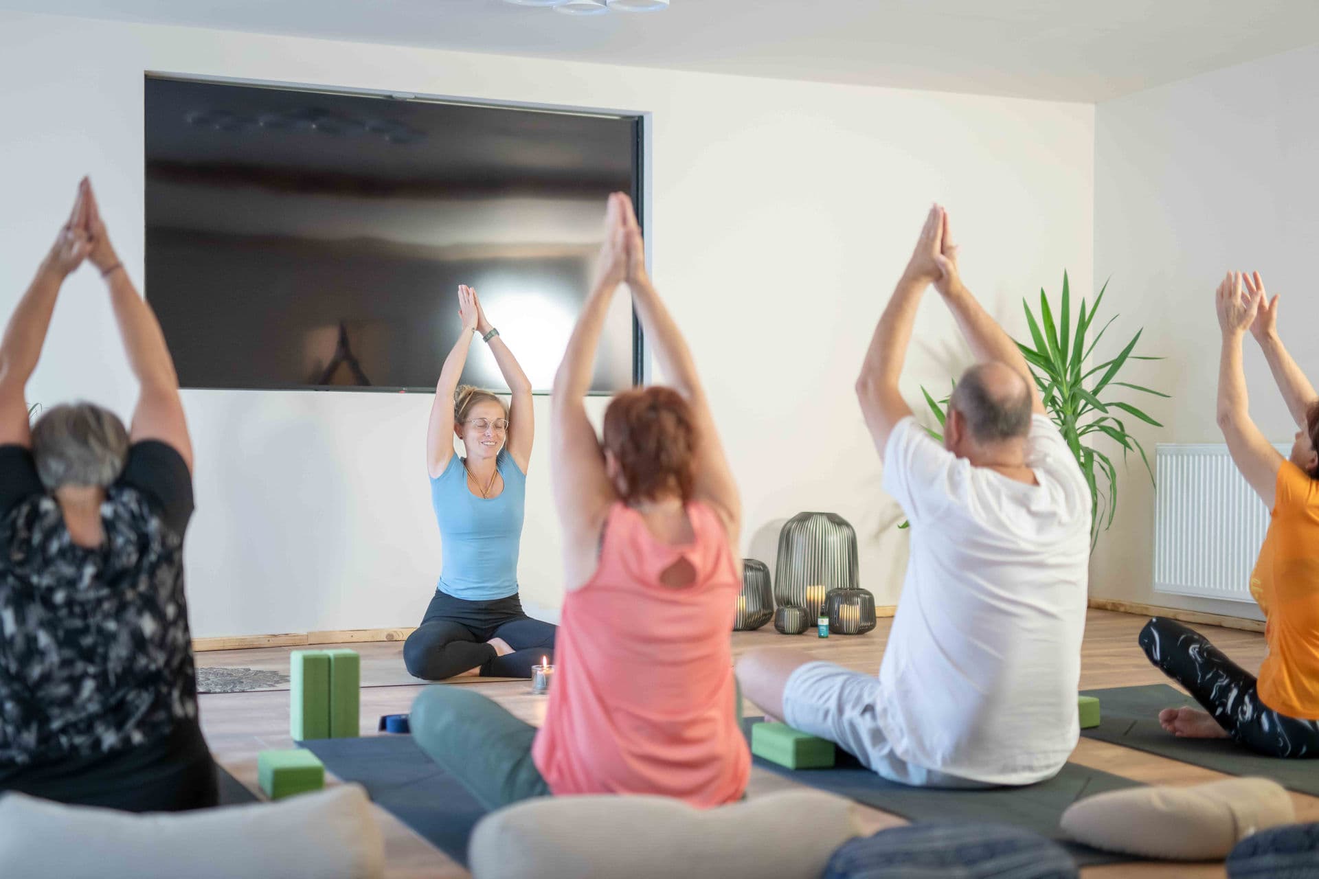 Menschen in einem hellen Raum praktizieren Yoga mit erhobenen Händen unter Anleitung einer Trainerin.