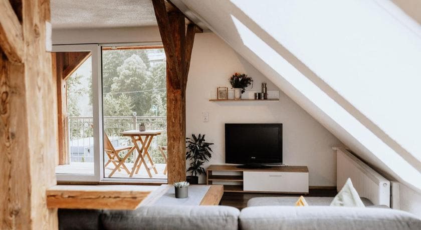 Helles Dachgeschoss-Wohnzimmer mit rustikalen Holzbalken, Fernseher und Balkon mit Blick auf grüne Bäume.