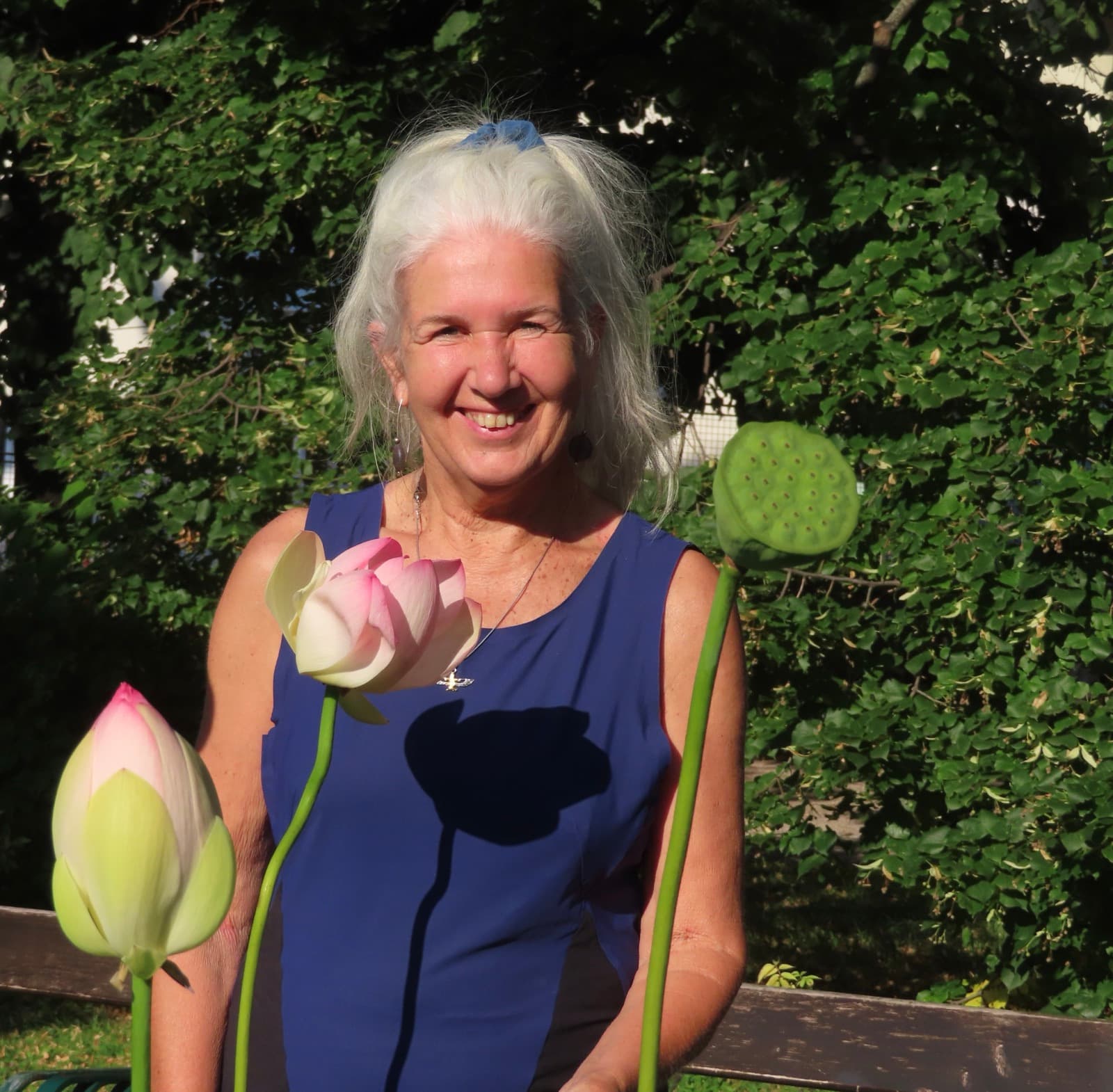 Eine lächelnde Frau mit weißen Haaren posiert hinter Lotusblüten und einer Samenkapsel vor grünem Laub.