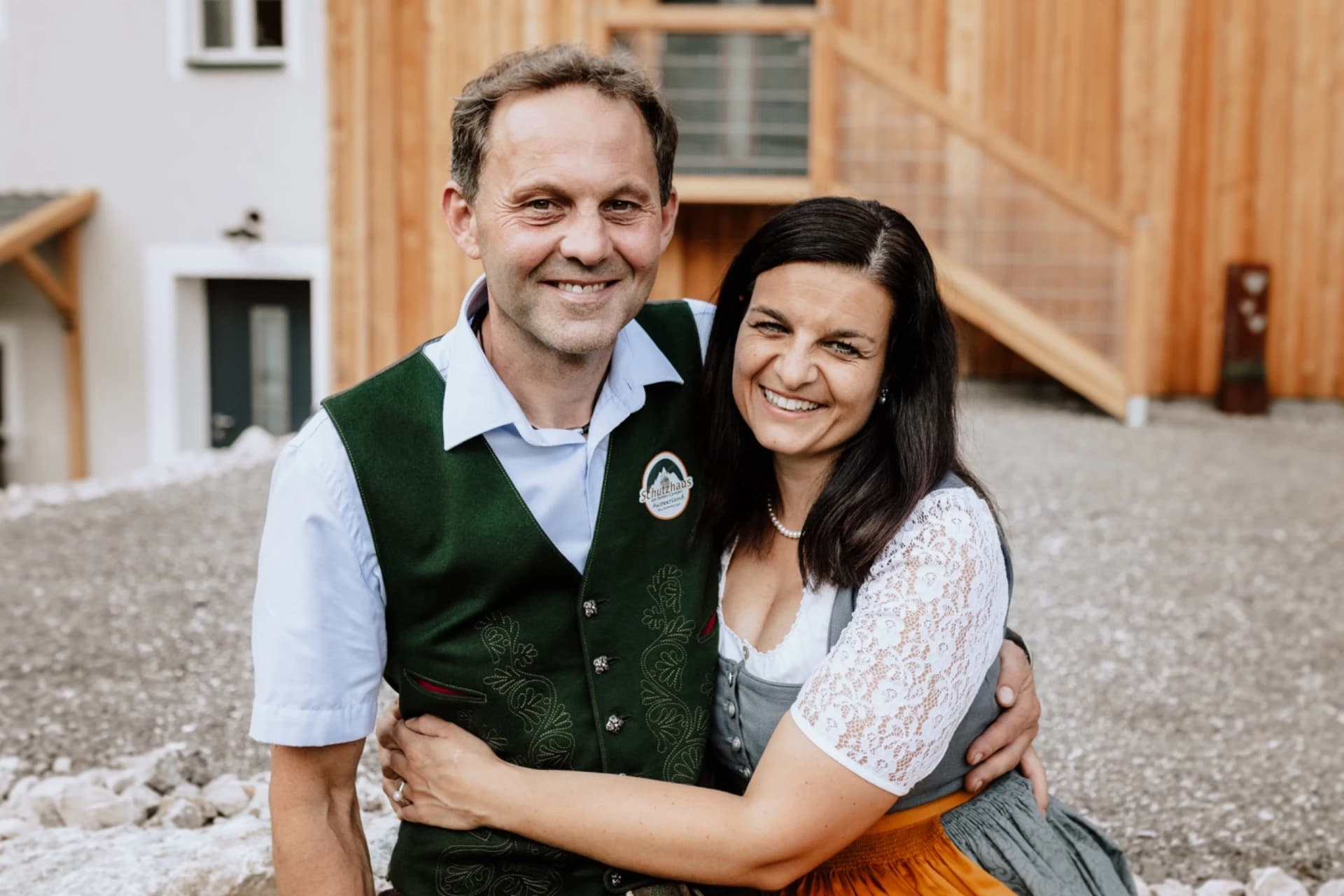 Ein lächelndes Paar in traditioneller Tracht posiert glücklich vor einem rustikalen Holzgebäude.