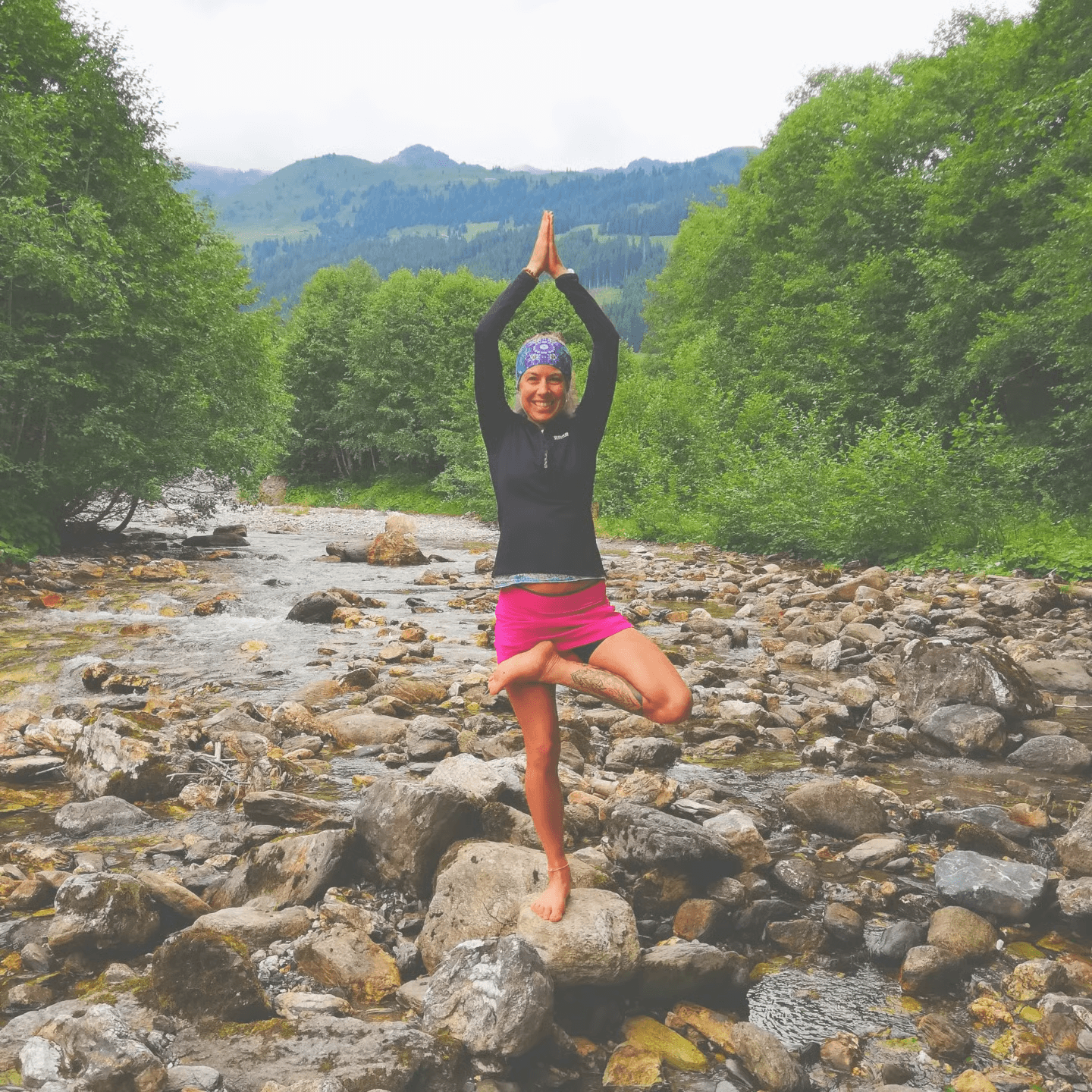 Lächelnde Frau in Yoga-Pose auf einem Stein im felsigen Fluss vor grünen Bergen.