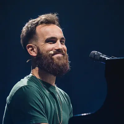 Lächelnder bärtiger Mann im grünen T-Shirt am Klavier vor einem Mikrofon auf dunkler Bühne.