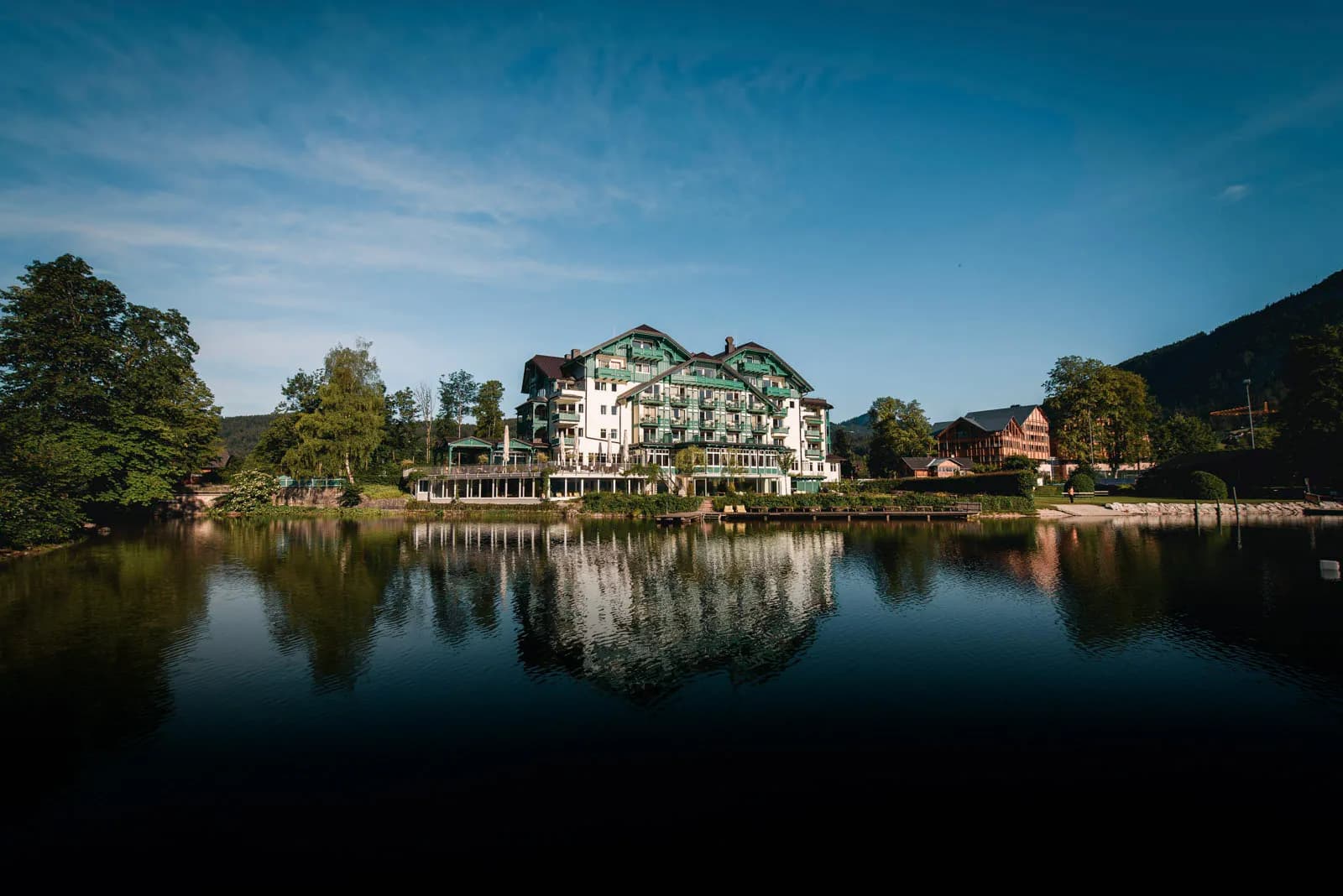 Großes Hotel mit grünen Balkonen spiegelt sich in einem ruhigen See unter klarem blauem Himmel.